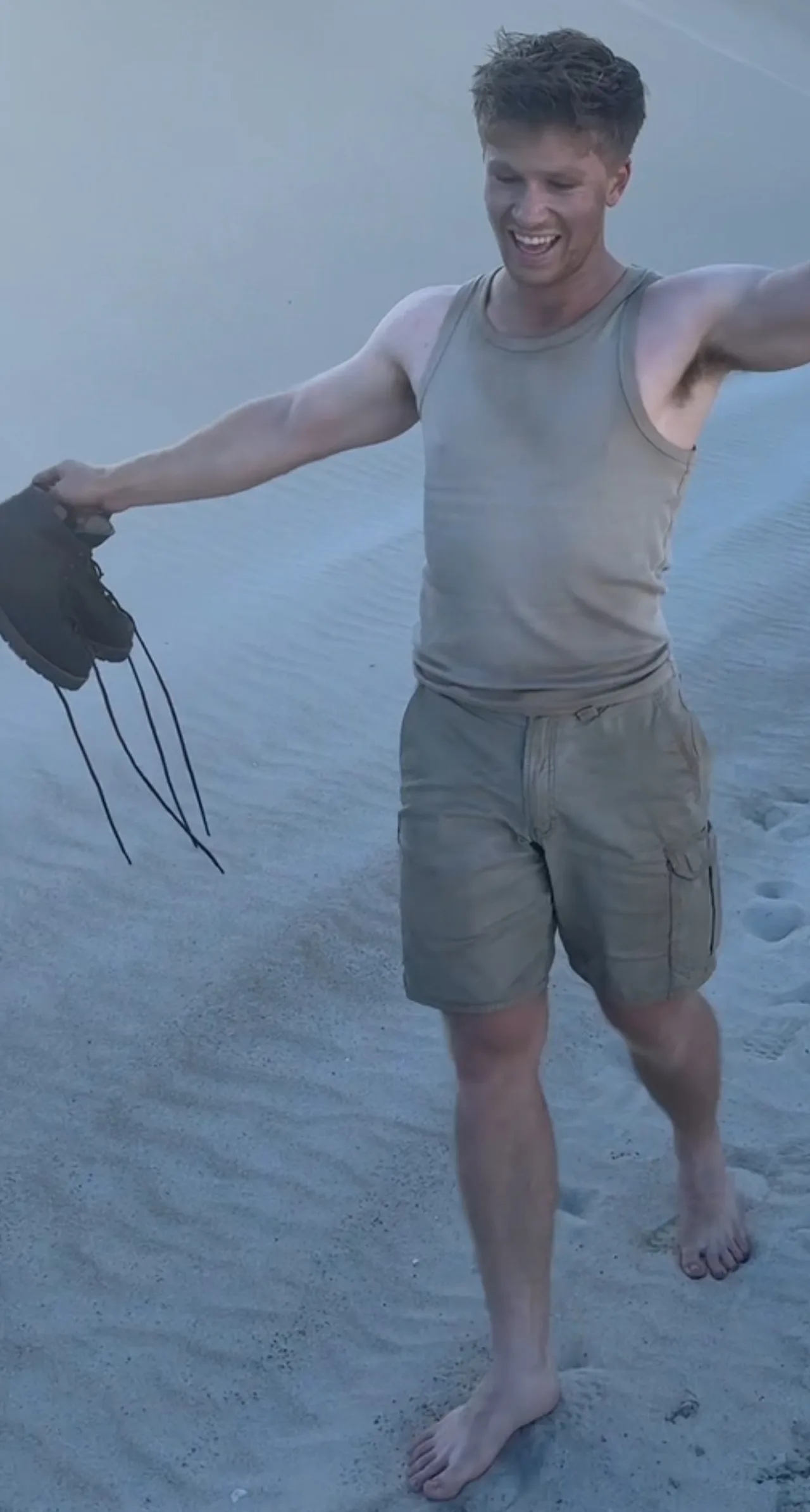 Robert Irwin grinning Barfoot på Sand i en Tank Top og Shorts