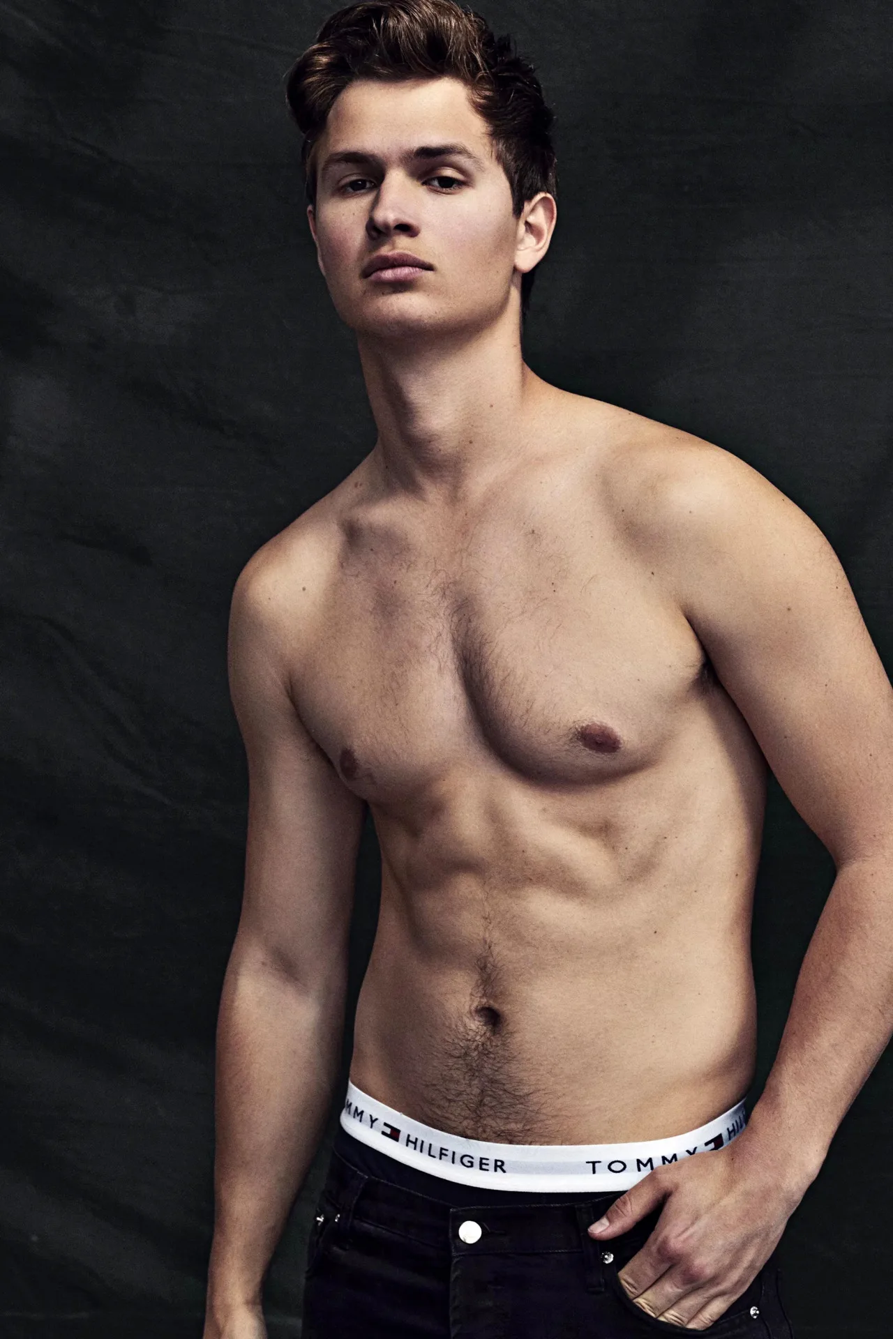 Ansel Elgort shirtless with defined abs, wearing Tommy Hilfiger underwear and dark jeans against a black backdrop