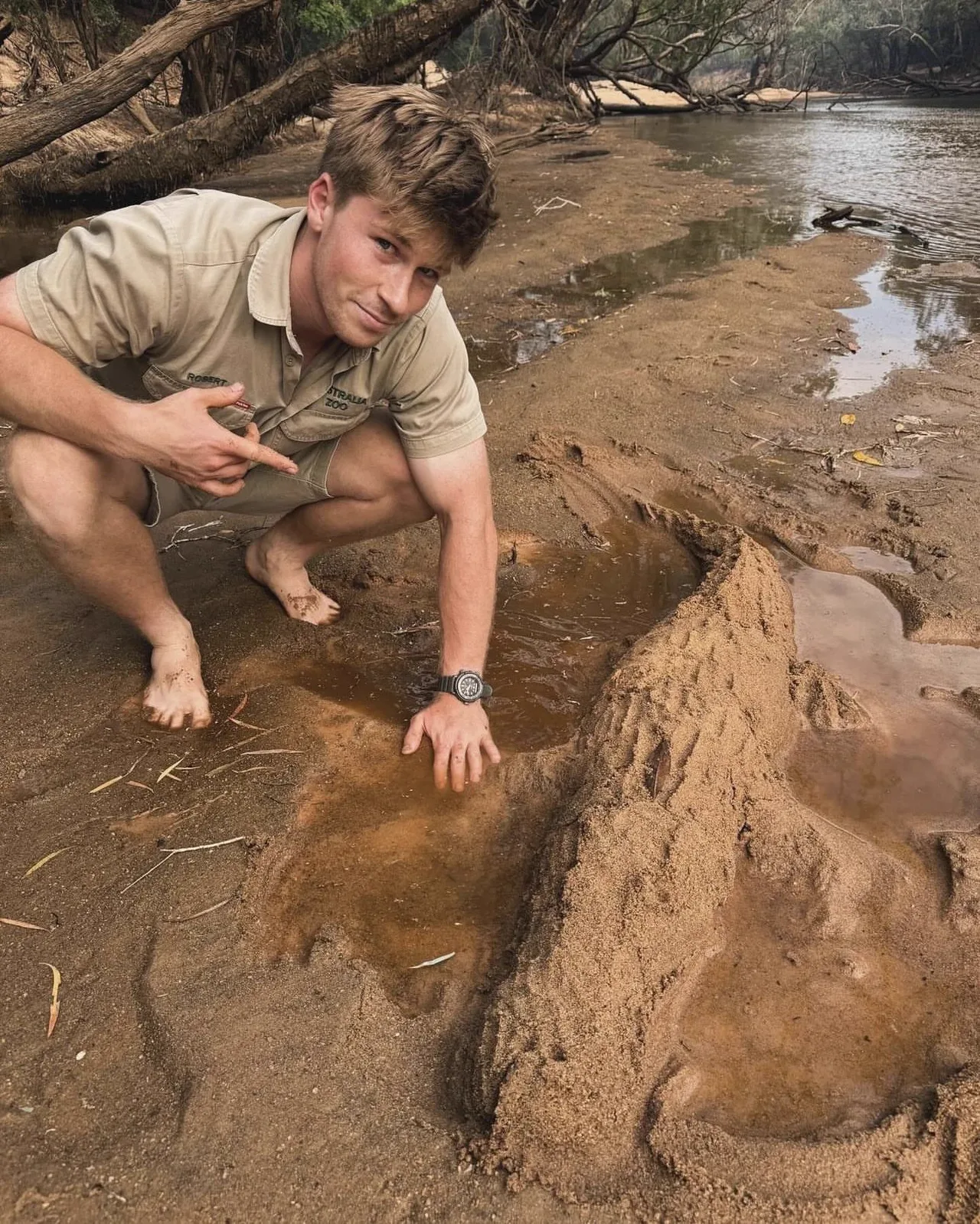 Robert Irwin shirtless in khaki shorts crouching barefoot by a muddy riverbank
