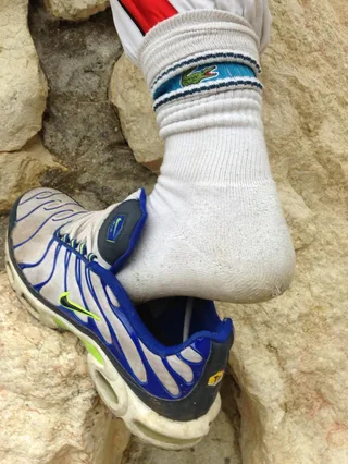 Um cara de meias de Lacoste e Nike Pumas mostra seu pé contra a parede de pedra