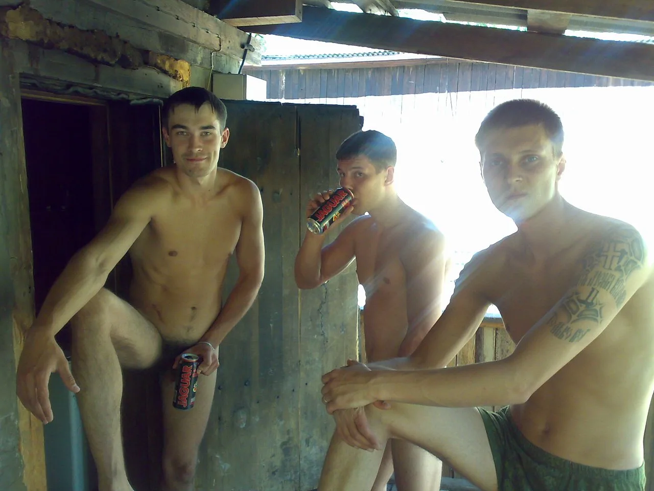 Drei hemdlose slawische Jungs in Armee-Shorts posieren mit Energy-Drinks unter rustikalem Holz