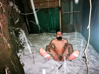 Um cara nu preso na neve com uma mordaça e correntes no tornozelo, totalmente exposto