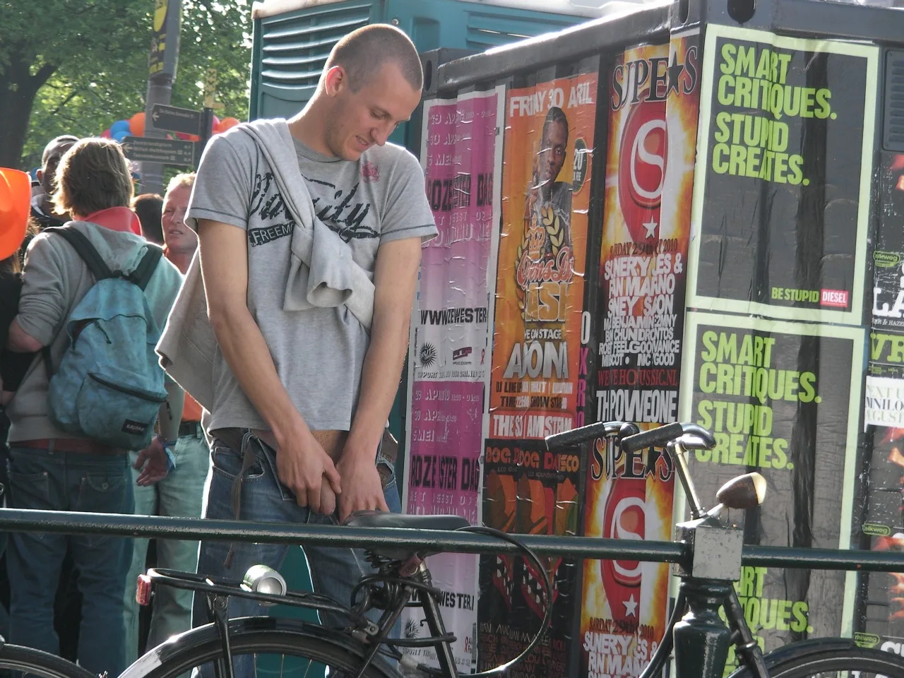 Guy i grå tee og jeans justerer skridtet med hænder, mens stående nær cykel og plakater