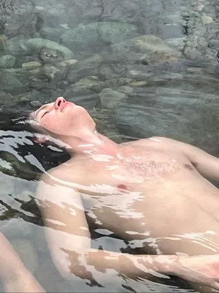 Ansel Elgort flutuando sem camisa em água limpa, peito nu brilhando sob luz natural