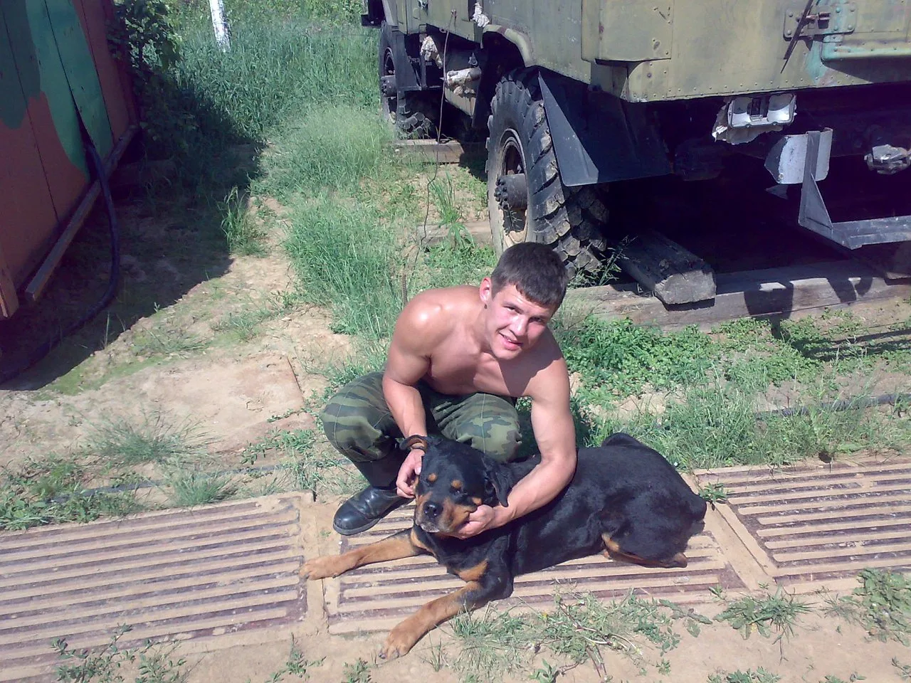 Rapaz de estilo militar sem camisa com calças de camuflagem. Animais Rottweiler ao lado de um caminhão blindado
