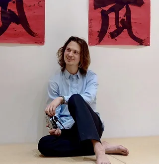 Ansel Elgort em camisa azul clara, descalço em tatami, segurando uma câmera vintage com um sorriso brincalhão