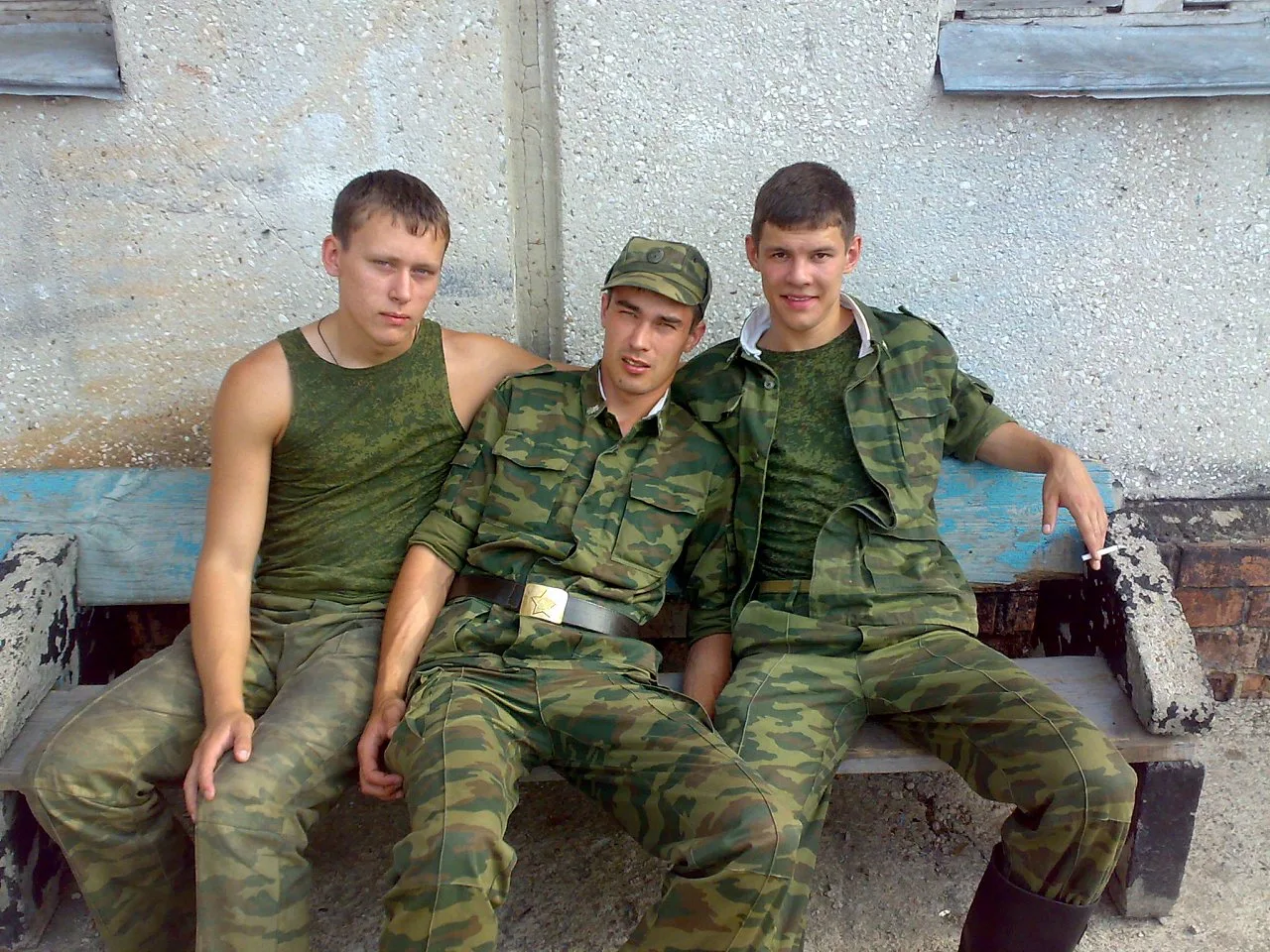 Três caras de estilo militar com roupas de camuflagem com peito nu num banco com um fumando