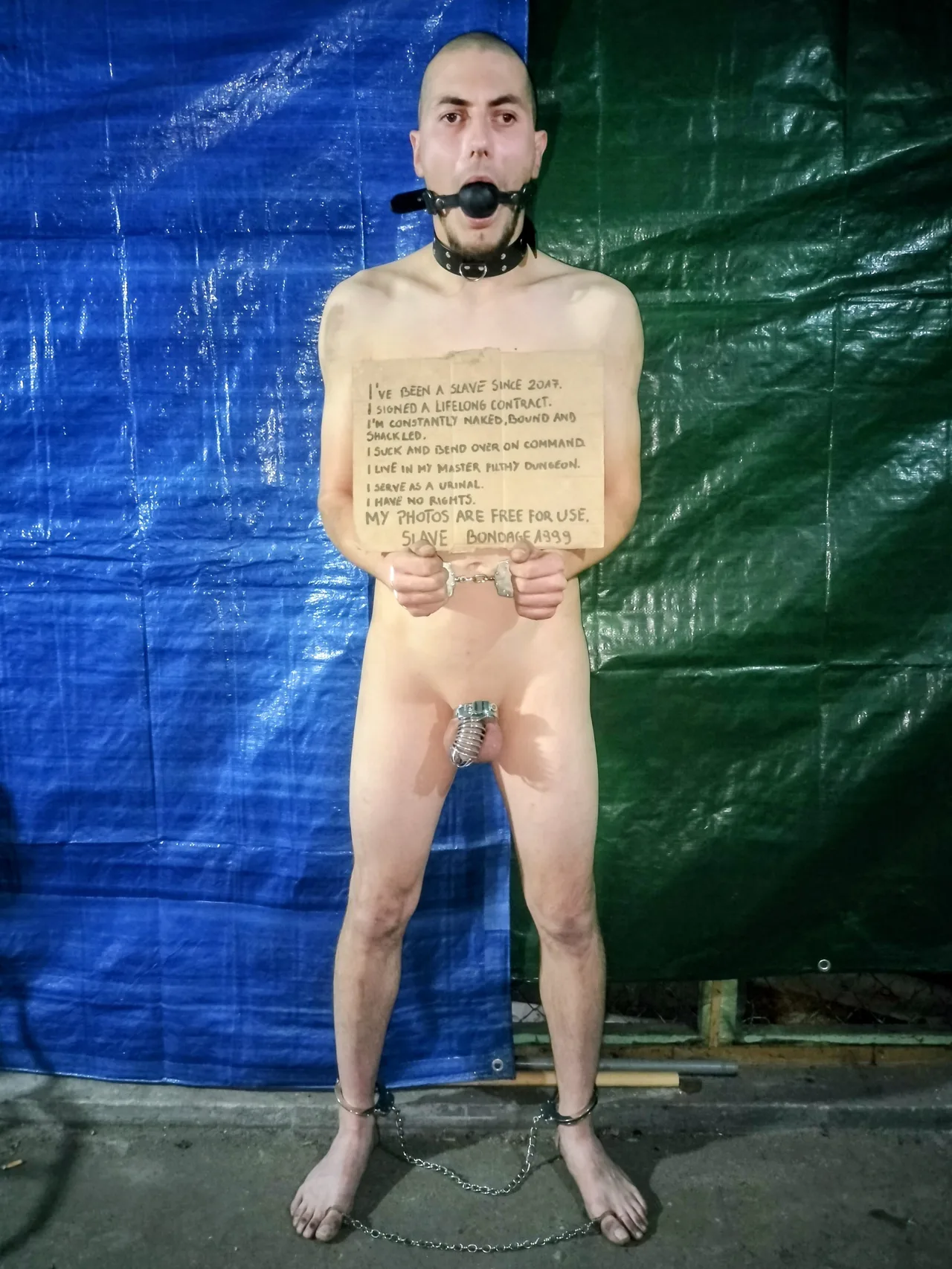 Hardcore slave fyr i fuld bondage med pik bur, bold gag, og kæder holder et skrifte tegn