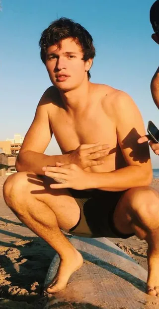 Ansel Elgort sem camisa na praia, agachado com pele beijada pelo sol e construção atlética em costa arenosa