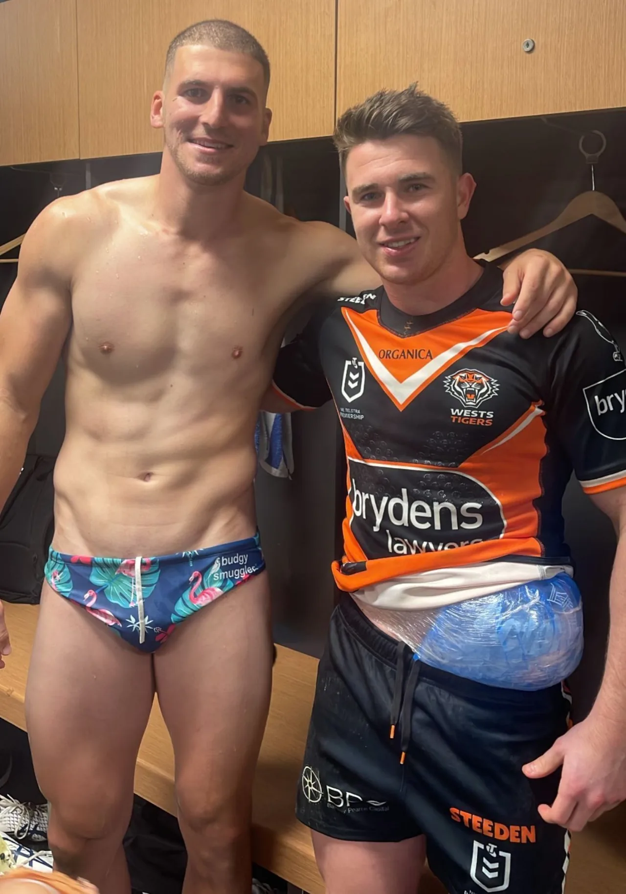 Due ragazzi ripped in spogliatoio, uno senza camicia in bauli di fenicottero, altro in kit rugby con pacchetto di ghiaccio