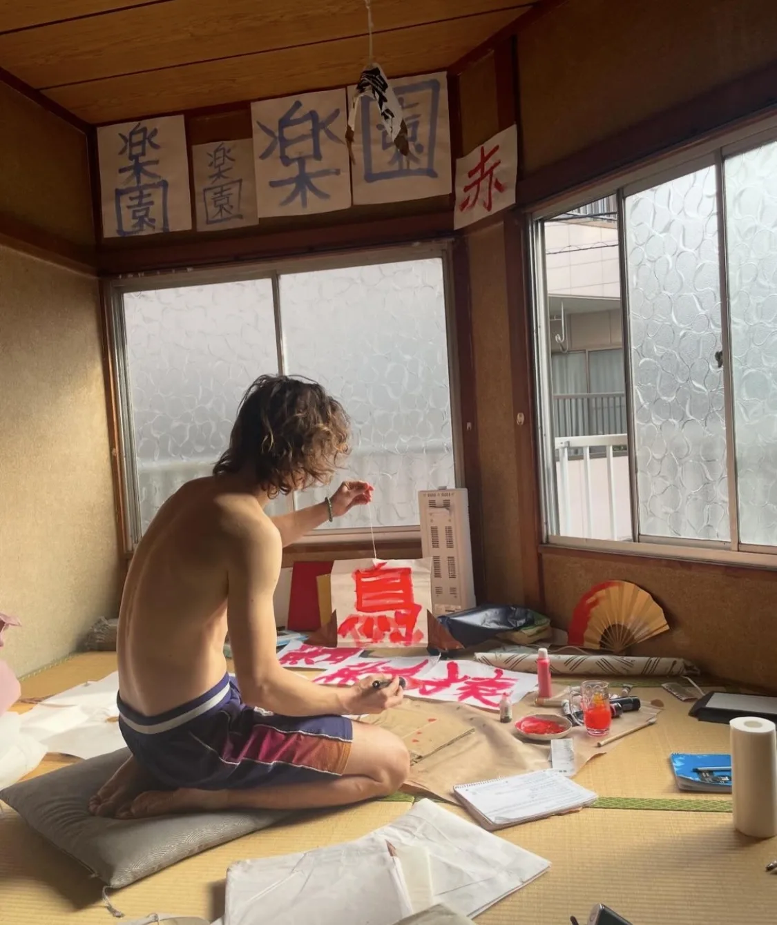Shirtless Ansel Elgort kneels on tatami, painting bold red kanji in a sunlit traditional room