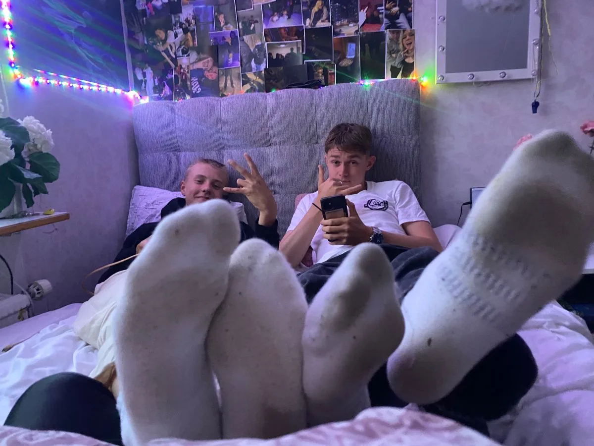 Dois rapazes relaxando na cama com os pés para cima, um telefone rodando, vibe aconchegante