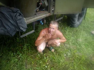 Um rapaz de estilo militar sem camisa agachado descalço na grama ao lado do caminhão, tudo natural e suado