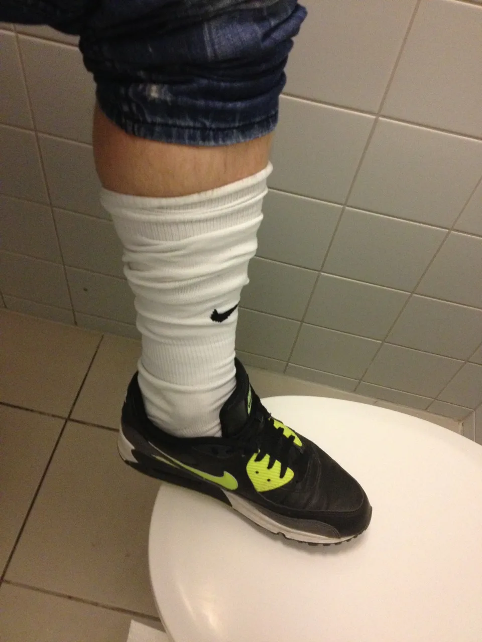 Cara de meias brancas e Nikes pretos empoleirados no banheiro, jeans enrolados alto para o foco dos pés