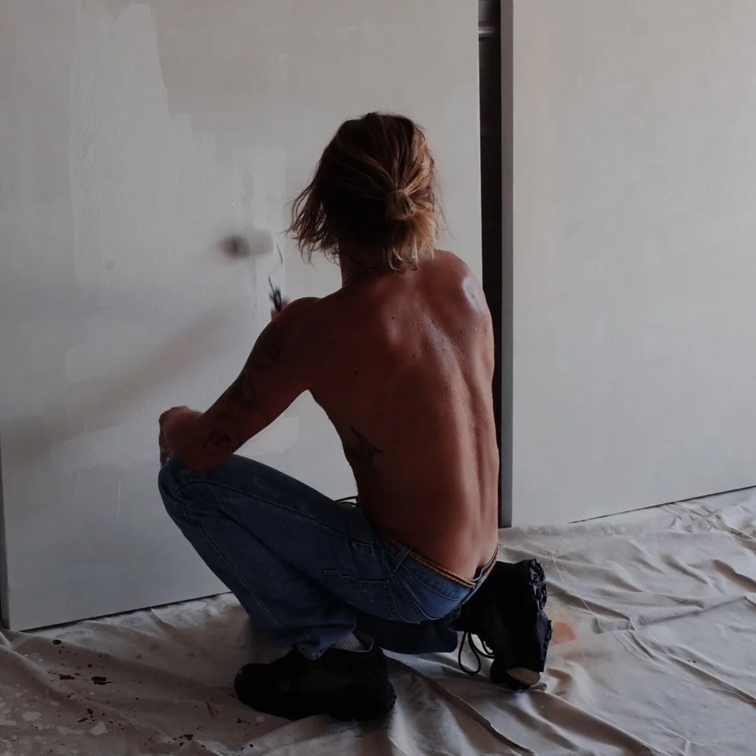 Hart Denton shirtless and squatting while painting a wall in jeans