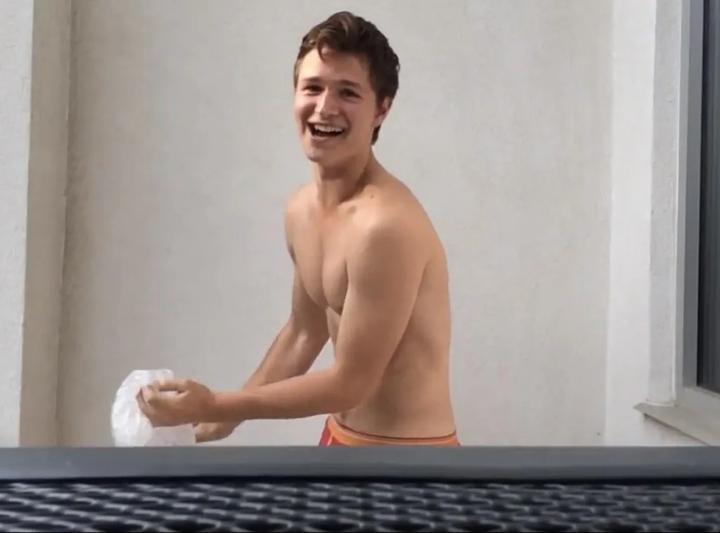 Ansel Elgort shirtless and smiling, bare torso gleaming in daylight, athletic build on display