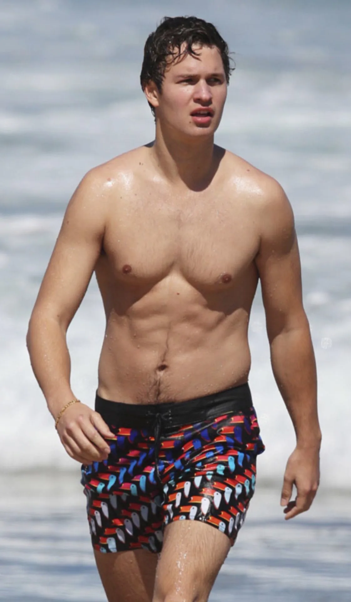 Ansel Elgort glatløs på stranden, våd hud glinsende, rokkende farverige svømmetunke med toucan print
