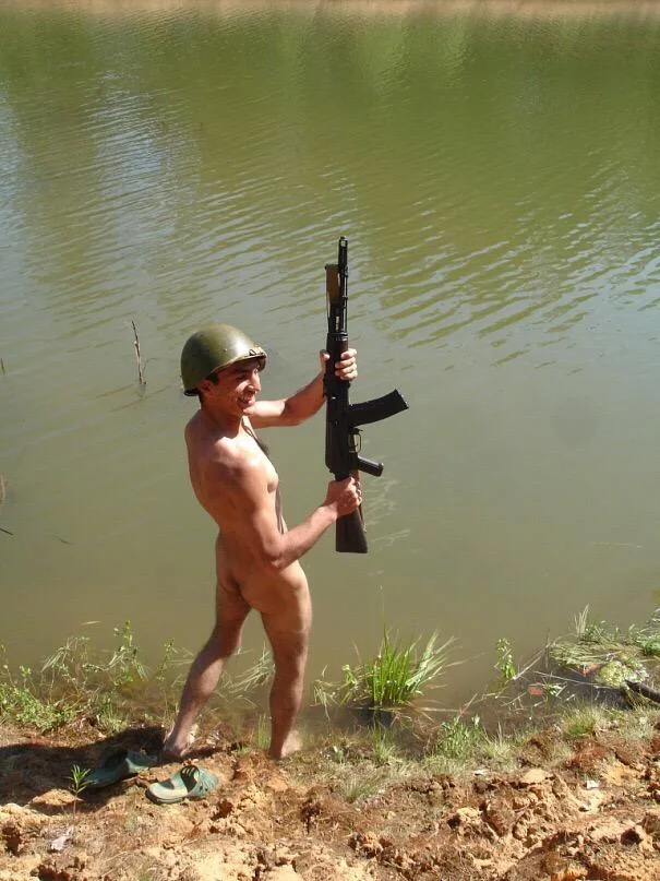 Nøgen soldat dreng griner med riffel ved mudret dam, hjelm på, støvler off
