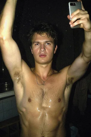 Ansel Elgort selfie sem camisa com pele molhada, braços levantados, e cabelo no peito brilhando sob luz fraca