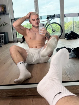 Um cara loiro de meias brancas tira selfie de espelho, pernas abertas, mostrando pés e abdominais tonificados
