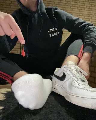 Guy in hoodie points at his socked foot beside a sneaker, casual foot-fetish vibe