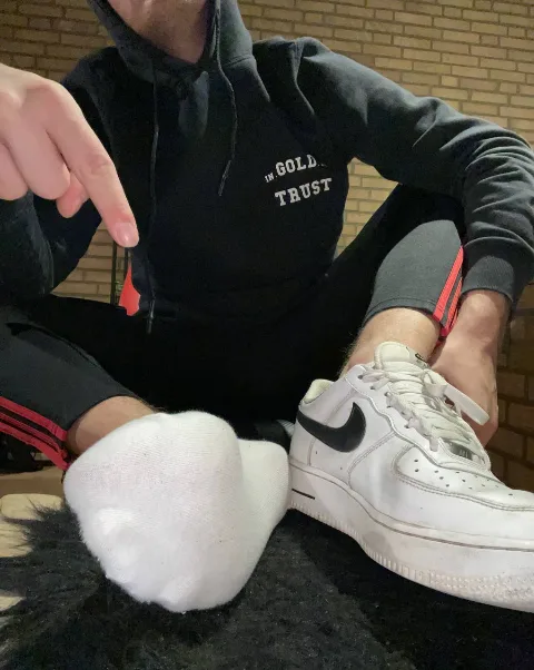 Guy in hoodie points at his socked foot beside a sneaker, casual foot-fetish vibe