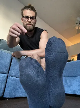 Bearded guy in black tank top points while showing off his socked feet on blue couch