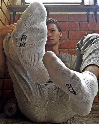 Lazy lad in grey socks flaunts soles while lounging shirtless on brick floor