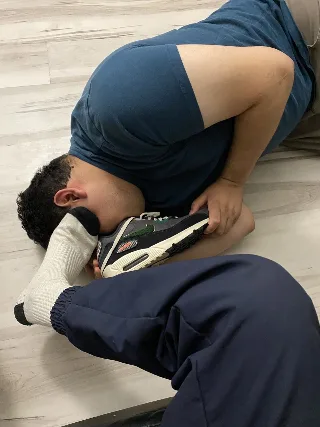 Guy in blue tee and sweatpants worships sneaker-clad foot, socked toes pressed to his face