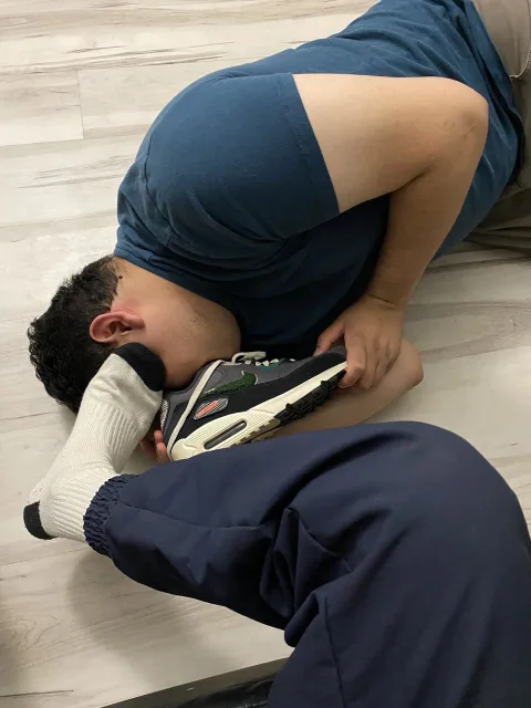 Guy in blue tee and sweatpants worships sneaker-clad foot, socked toes pressed to his face