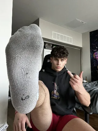 Guy in red shorts flashes his socked sole while flipping the bird, hoodie on, bedroom vibe