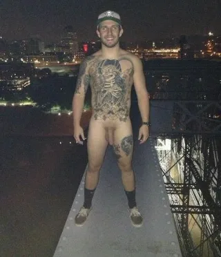 Tattooed lad in cap stands naked on bridge at night, city lights behind his hard cock