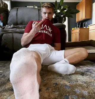 Sock-clad guy lounging with one foot forward, red tee and white shorts, casual living room vibe