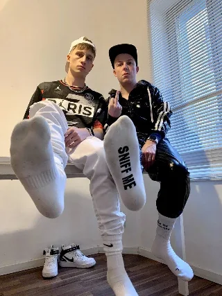 Two lads flaunt socked soles with cheeky foot text, one giving the finger in shiny black gear