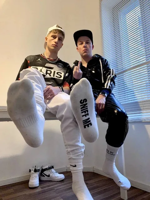 Two lads flaunt socked soles with cheeky foot text, one giving the finger in shiny black gear