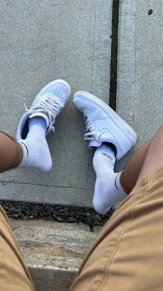 White sneaker-clad feet in ribbed socks, legs spread on concrete, casual street vibe