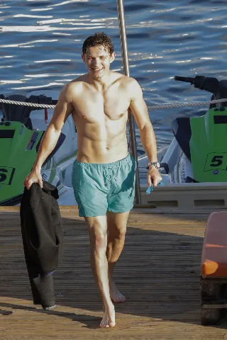 Tom Holland shirtless on dock, smiling with swim trunks and wetsuit in hand by jet skis