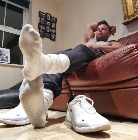 Lazy lad lounging with socked soles up, one sneaker kicked off, relaxed at home