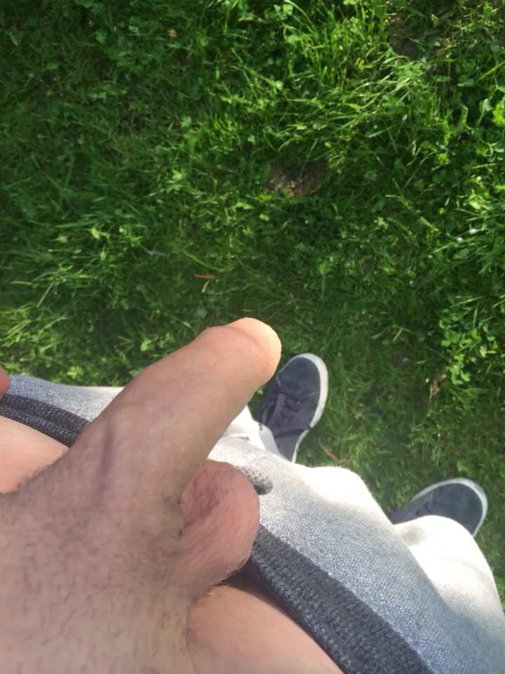 Homem de moletom cinza puxa a cintura, polegar pra cima, exibindo o pau ao ar livre
