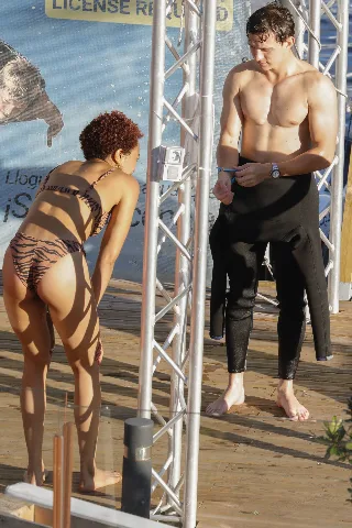Tom Holland shirtless on dock, adjusting wetsuit with toned abs and watch, sunlit beach vibe