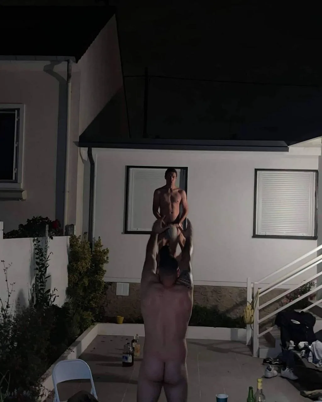 Two naked lads partying hard outdoors, one lifting the other for a cheeky rooftop pose