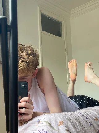 Curly blond lad in tank top snaps mirror selfie with bare feet up on floral bed
