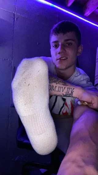 Sock-clad foot flexed close-up with tattooed lad in purple glow, shirtless and smirking