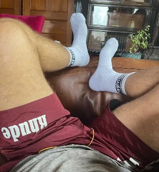 Lazy lad in maroon shorts and white socks kicks back on leather couch, feet up for foot fetish fans