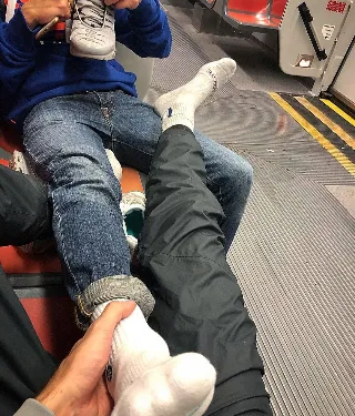 Guy in blue hoodie and jeans holds sneaker while pal massages his socked foot on subway