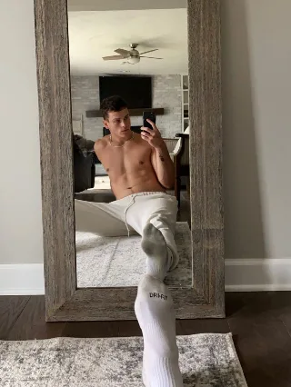 Shirtless lad in white socks takes mirror selfie, one leg kicked up showing socked foot