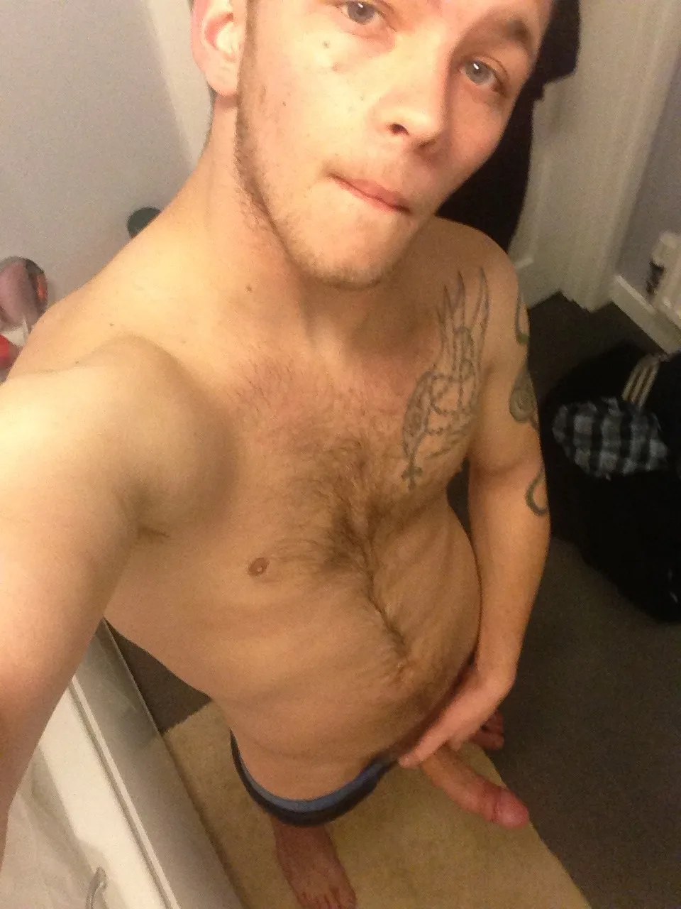 Garoto scally sem camisa, com tatuagens no peito e barriga peluda, posando com o pau duro na mão