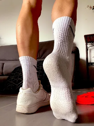 Muscular guy in white socks and sneakers flexes calves, one foot bare on floor, rear view