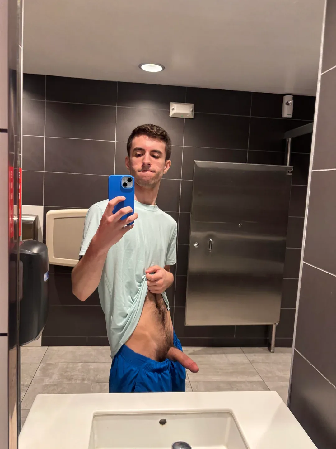 Garoto no espelho do banheiro tirando selfie e levantando a camisa para mostrar o pau duro e os pelos pubianos
