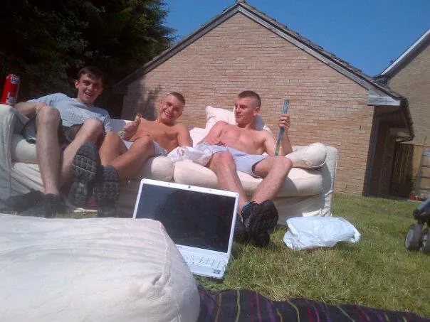 Three lads in shorts lounging shirtless on garden sofa with beer and laptop